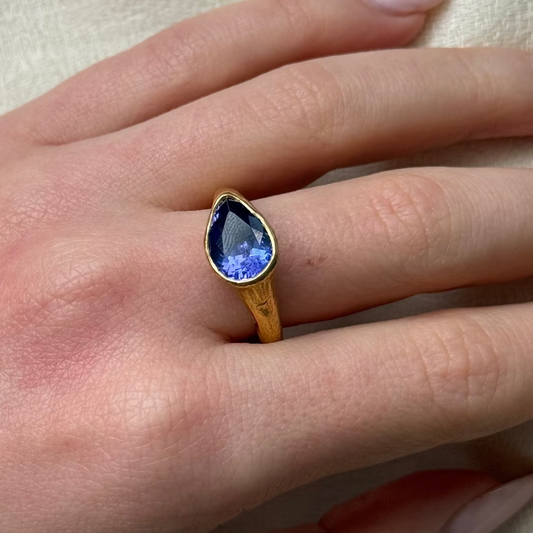 Hand wearing a 22ct gold ring with a blue gemstone on a neutral background