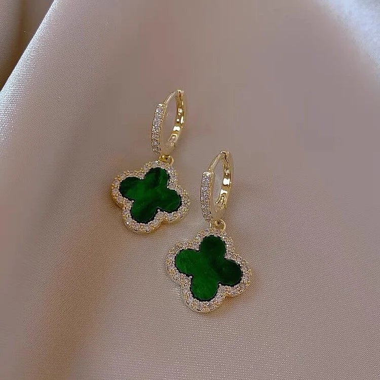 Gold earrings with green clover-shaped stones on a beige fabric background
