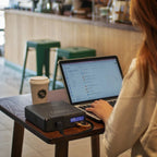 Person using a laptop with a portable power station on a table in a cafe.