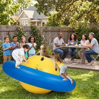 Children playing on a large blue and yellow wobble ball in a backyard with adults sitting at a table.