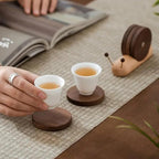 Two white teacups with wooden coasters on a textured surface, with a hand holding one of the teacups.