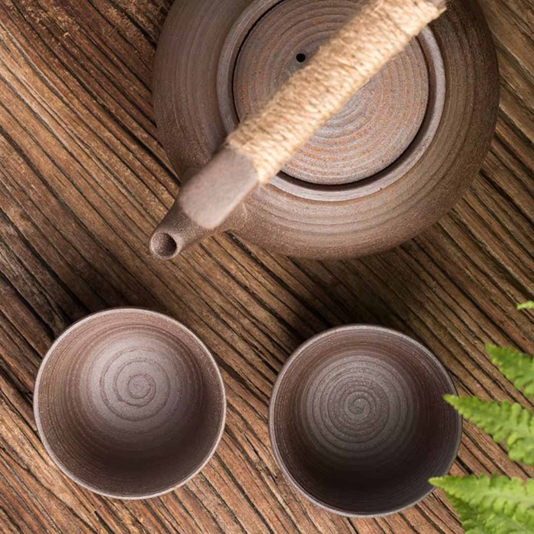 Ceramic teapot and two cups on a wooden surface with a plant in the corner.