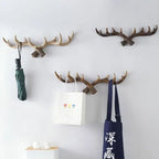 Three wall-mounted antler hooks with bags and an umbrella hanging on them against a white wall.
