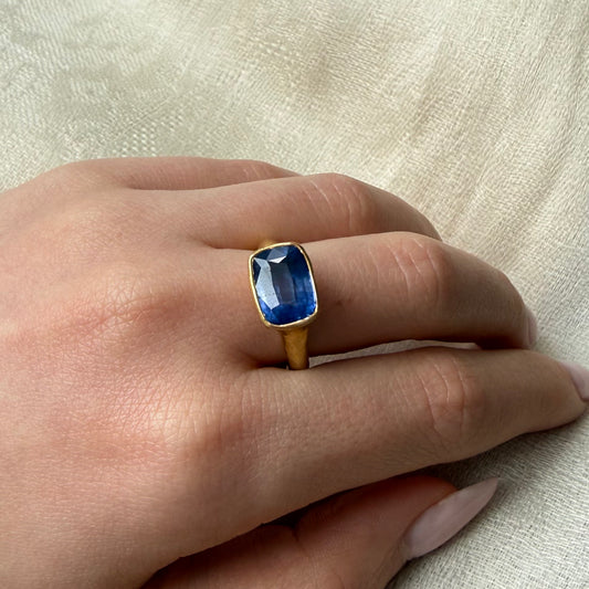 Hand wearing a 22ct gold ring with a blue Tourmaline gemstone on a light fabric background