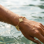 Hand wearing a gold bracelet with floral designs by a body of water.