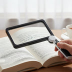 Hand holding a magnifying glass over an open book on a wooden surface.