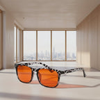 Sunglasses with orange lenses and black frame on a surface with a cityscape view through windows in the background.