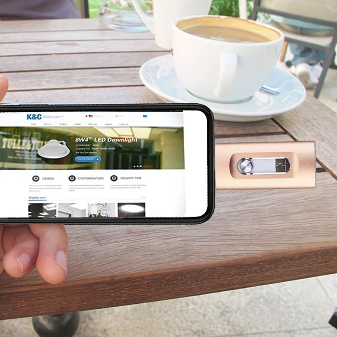 Person using a smartphone with K&C Lighting website on a wooden table with a coffee cup and saucer.