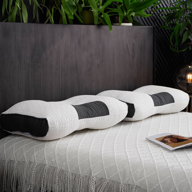 White and black wave-patterned body pillow on a bed with a dark headboard and plants in the background.