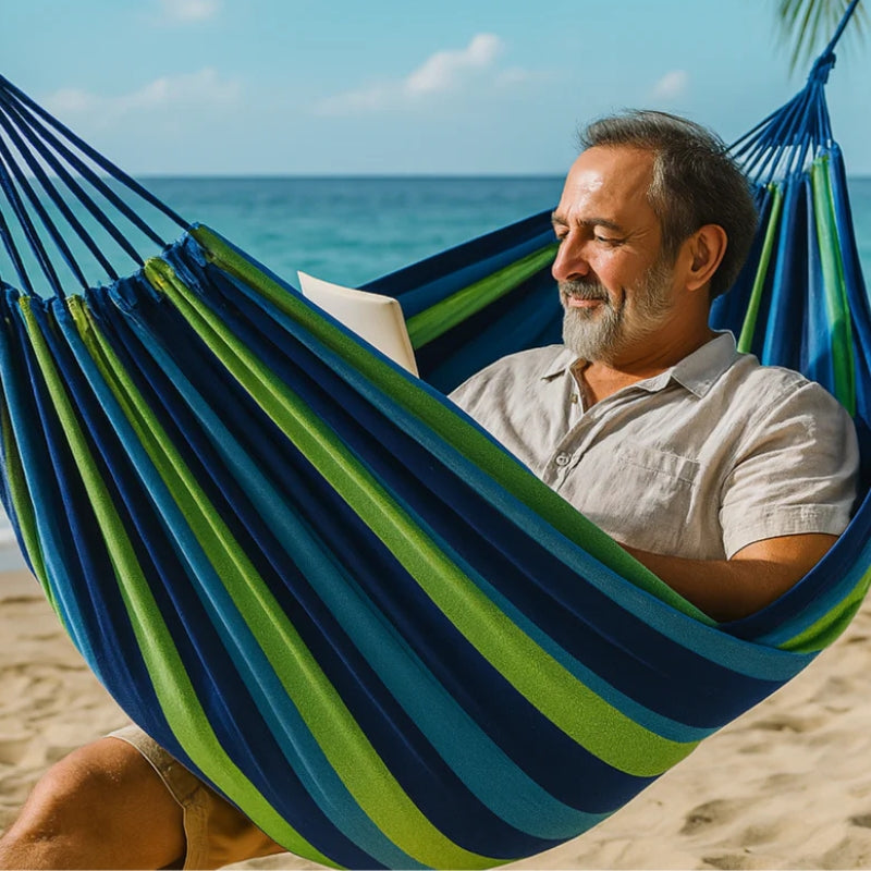 Man reading a book in a blue and green striped hammock on a beach.