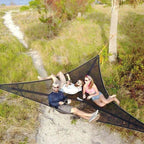 Three people relaxing in a large black hammock in a natural setting with grass and trees.