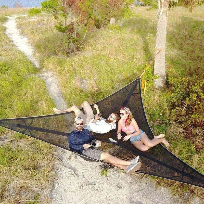 Three people relaxing in a large black hammock in a natural setting with grass and trees.