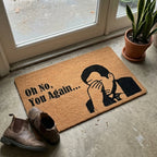 Doormat with a humorous design and text on a concrete floor next to a door.
