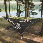 People relaxing in a large black hammock by a lake with trees in the background