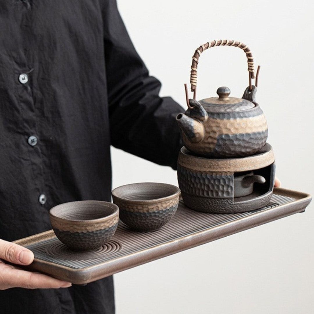 Person holding a tray with a ceramic teapot and two cups on a plain background