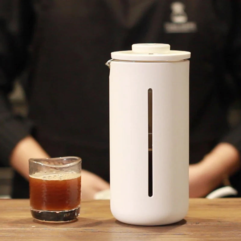 White coffee maker on a wooden table with a glass of coffee next to it.