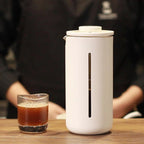 White coffee maker on a wooden table with a glass of coffee next to it.