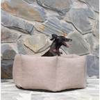 Dog sitting in a beige dog bed against a stone wall.