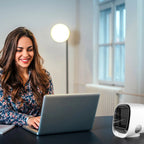 Woman using a laptop with a small air conditioner on a desk in a bright room.