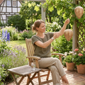Woman sitting in a garden holding a woven basket, surrounded by flowers and plants.