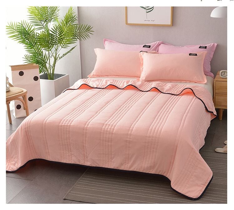 Pink bedspread and pillows on a bed in a bedroom setting with a plant and side tables.