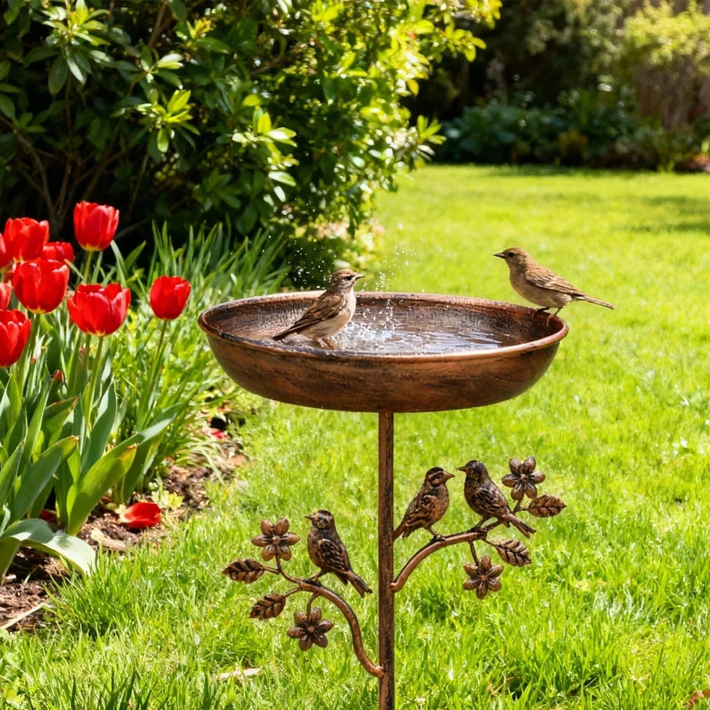 Bird bath with decorative birds on a stand in a garden with red tulips and green grass.