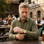 Man in a green shirt looking at his watch in an outdoor setting with people and buildings in the background.