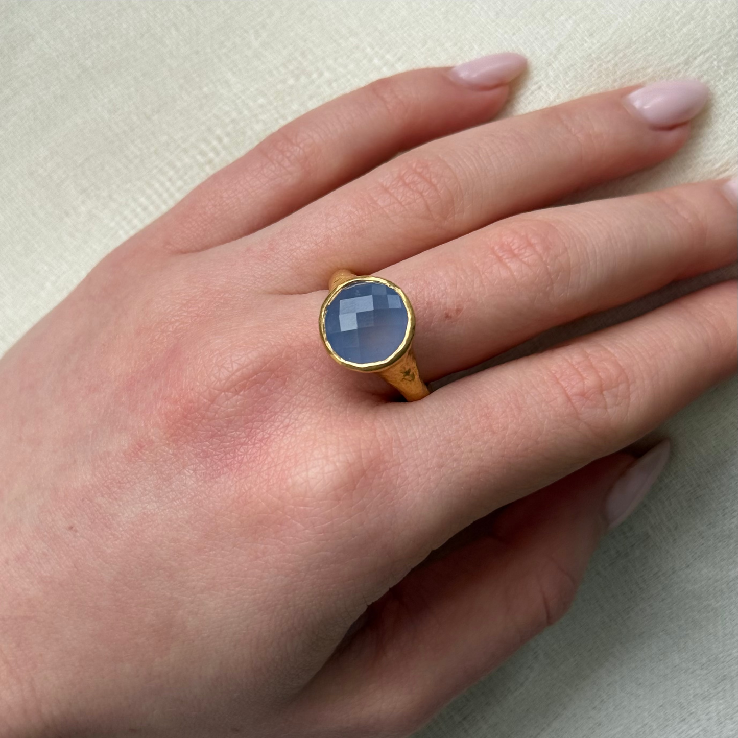 Hand wearing a 22ct gold ring with a blue stone on a neutral background
