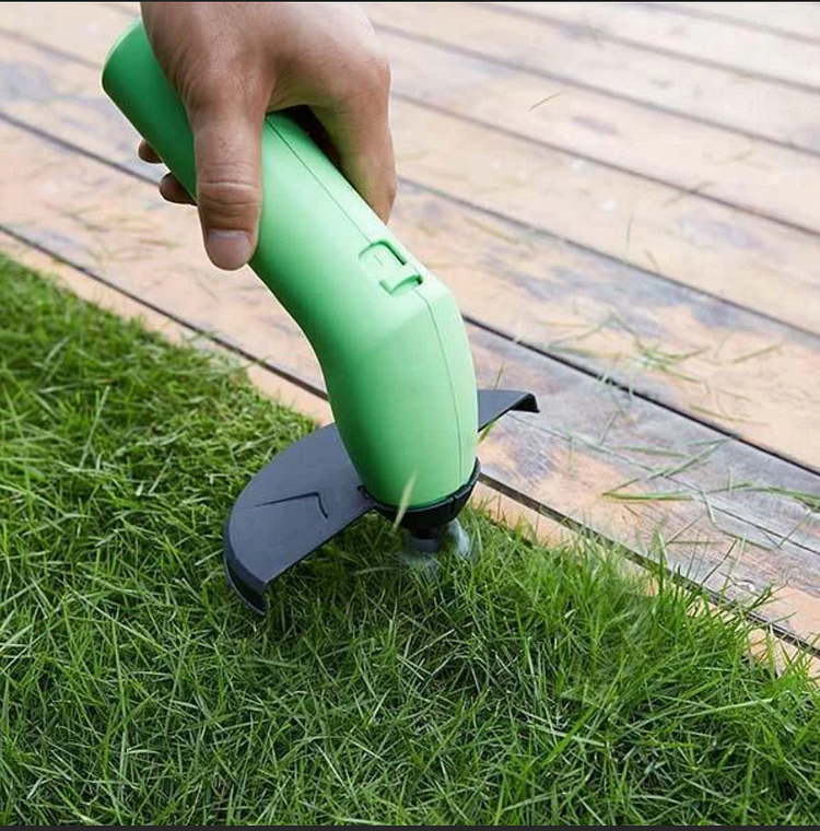 Hand using a green grass trimmer on grass next to a wooden deck.