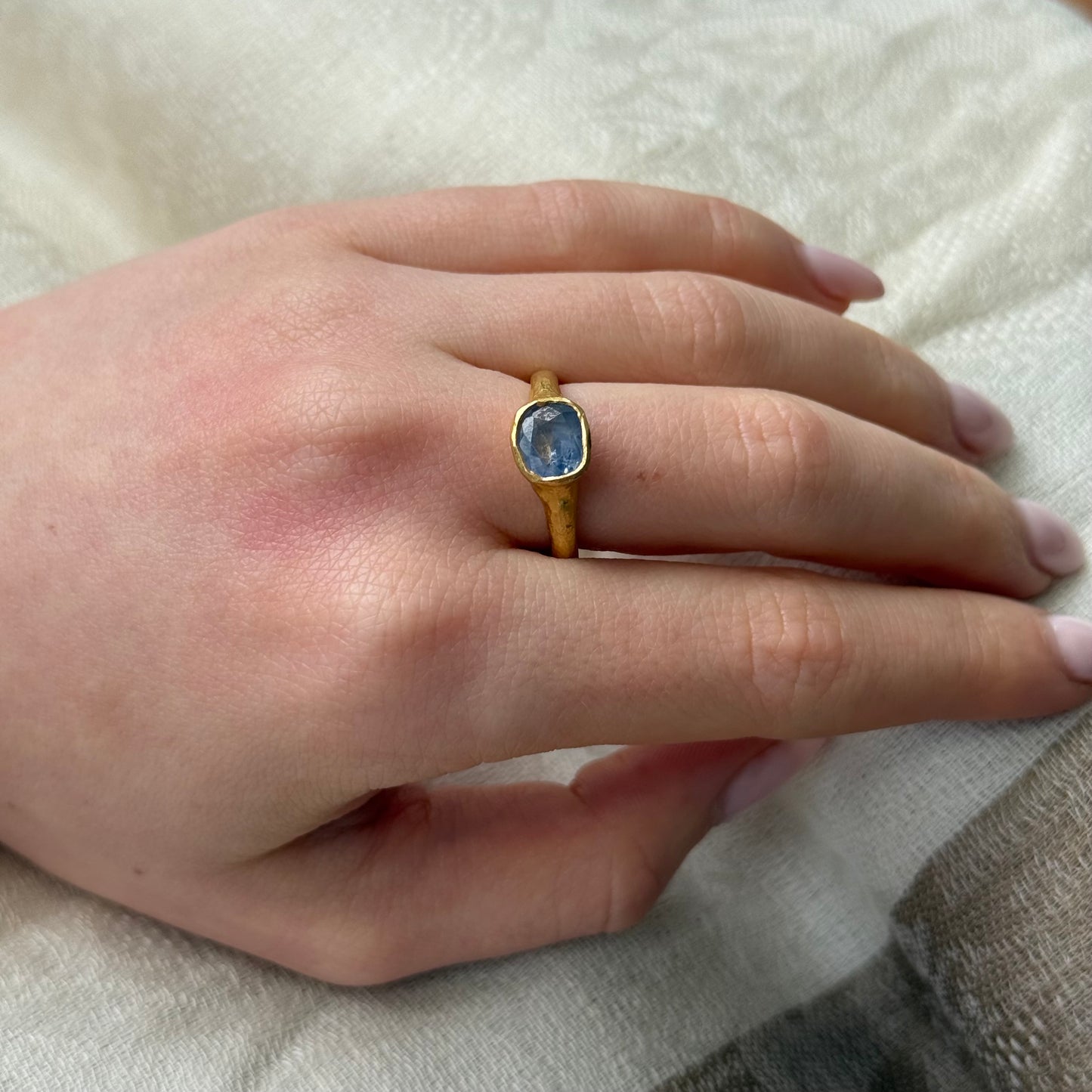 Hand wearing a 22ct gold ring with a blue sapphire gemstone on a light background