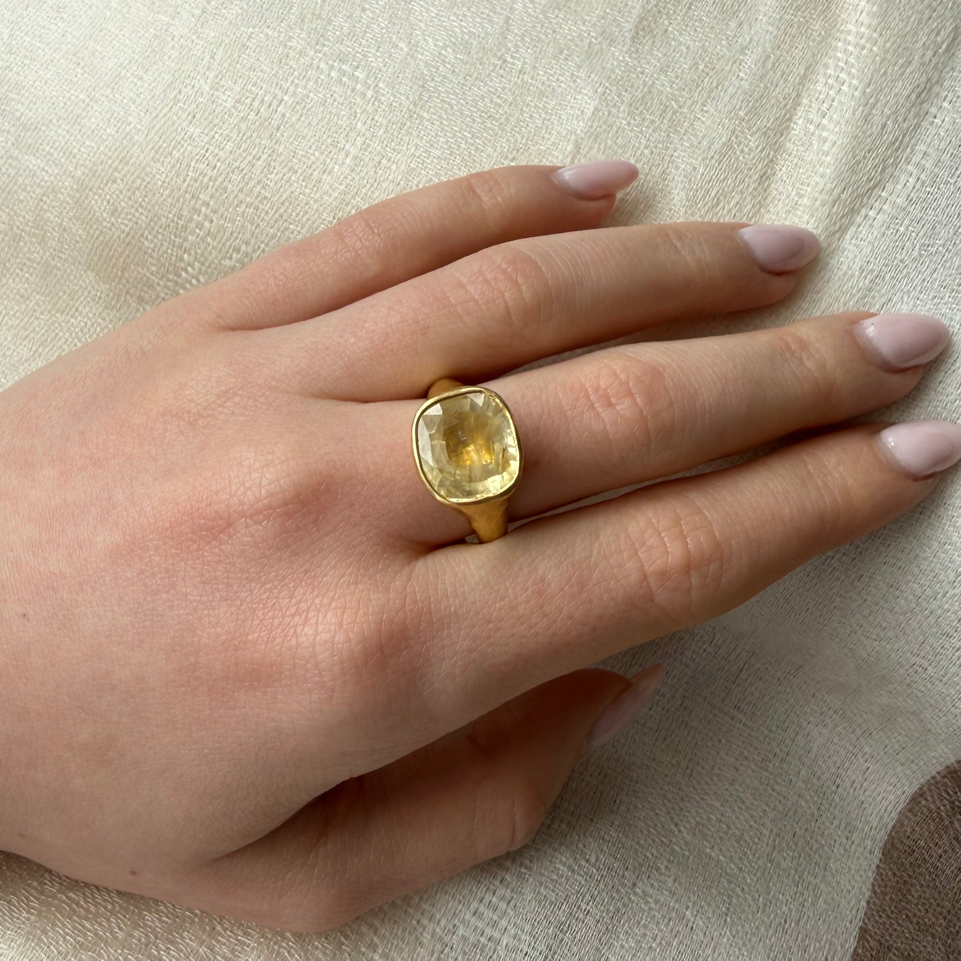 Hand wearing a 22ct gold ring with a yellow unheated natural sapphire 8 carat  gemstone on a beige fabric background