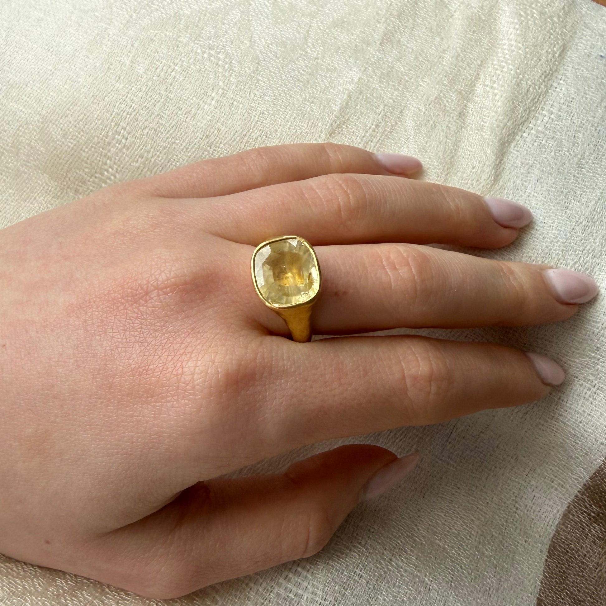 Hand wearing a 22ct gold ring with a large yellow sapphire natural unheated gemstone on a beige fabric background