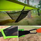 Close-up of a green hammock with mesh pocket and carabiner attachment, set against a natural outdoor background.