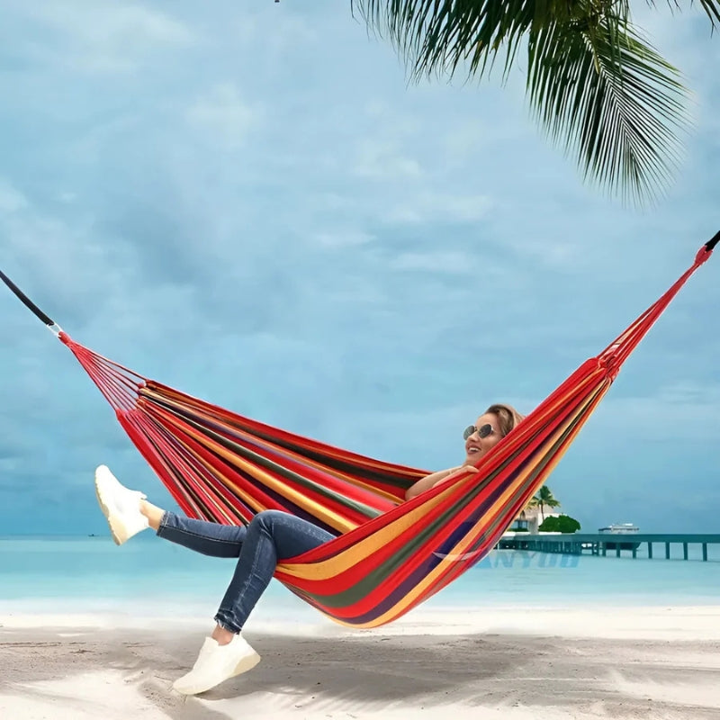 Person relaxing in a colorful hammock on a tropical beach with palm trees and clear blue sky.
