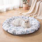 White cat lying on a patterned pet bed in a room with a plant and blanket.