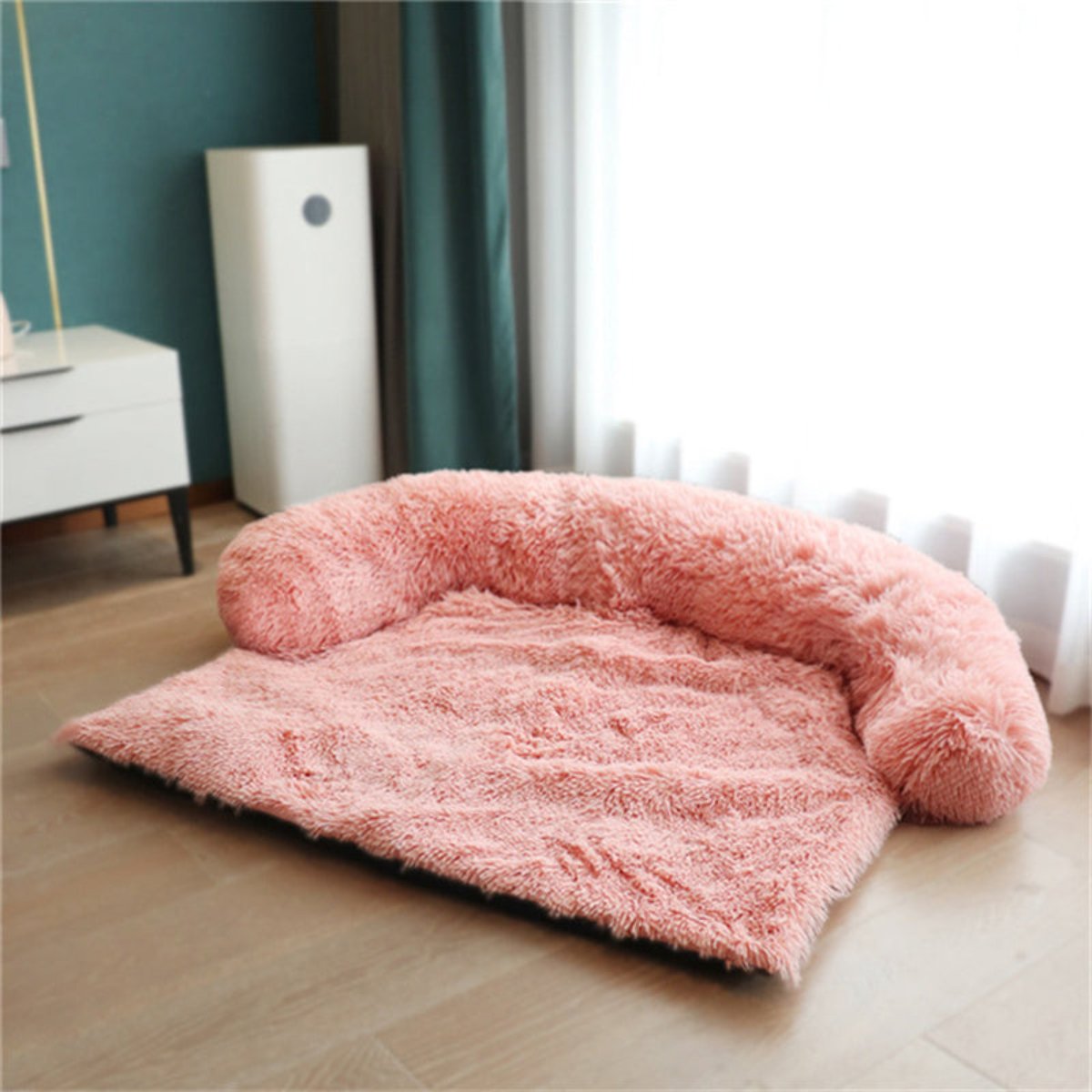 Pink fluffy pet bed on a wooden floor with a teal wall and white radiator in the background.