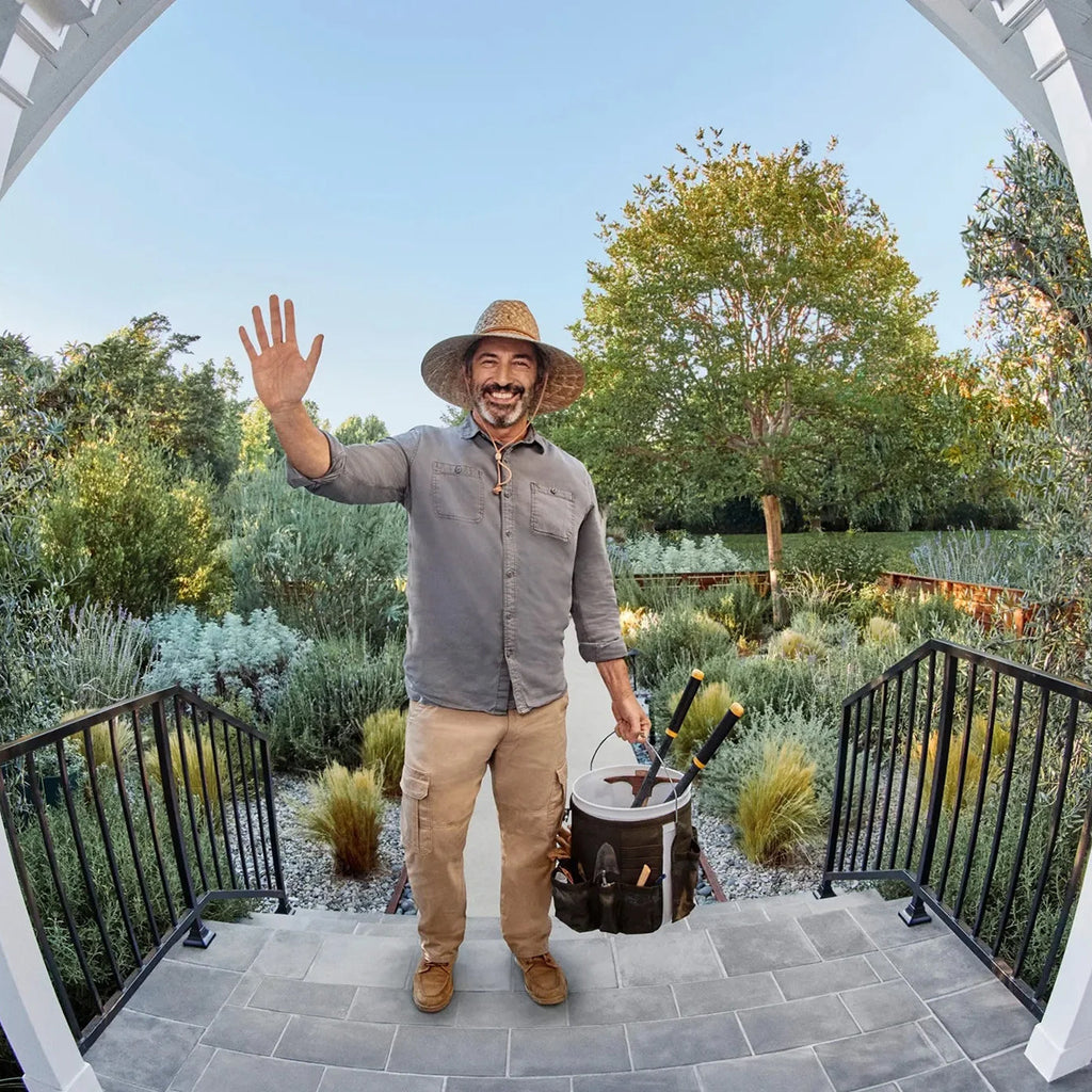 Man in a garden setting with tools, waving at the camera