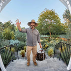 Man in a garden setting with tools, waving at the camera